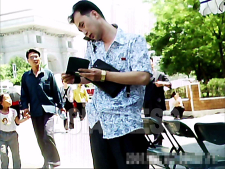 手帳をめくりながら携帯電話を使う男性。場所は平壌市の中心部。背後に凱旋門が見える。2011年6月牡丹峰区域、具光鎬（ク・グァンホ）記者撮影）