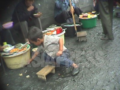 闇市場の露天食堂で客の食べこぼした米粒を拾う男児。90年代飢饉の際に全国どこでも見られた悲しい光景であった。（1998年10月江原道元山市　アン・チョル撮影）