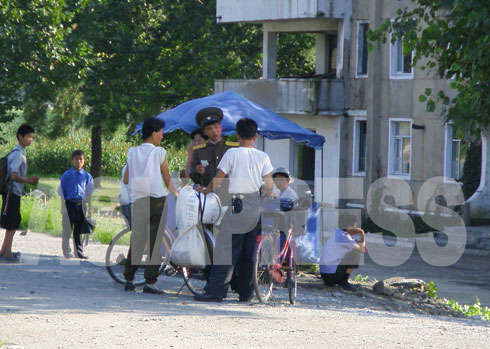 農村で仕入れた穀物を市場に持って行く途中、保安員（警察）の取り締まりに遭遇した二人。女性が自転車に乗ることは禁じられている。罰金だ。90年代に金正日総書記が女性が自転車に乗るのは見た目に悪いと言ったからだとか。その後うやむやになっていたのだが、最近またうるさくなったという。