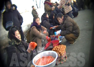 露天の食べ物屋で女性が腹ごしらえをしている。パンや餅などが売られている。写真手前のタライに入っている湯気の立った食べ物は何か不明。