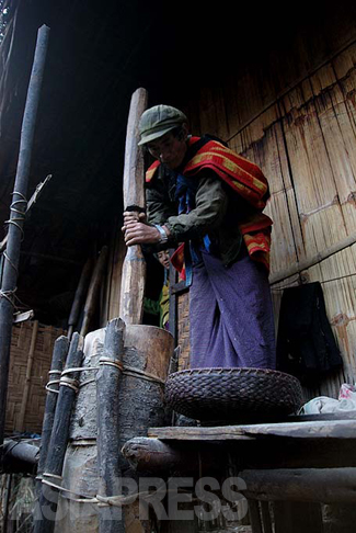 タストゥー村。子どもを背負い穀物を搗くラワン人男性。ビルマ最北の地にあって、中国の人民帽子を被っている。