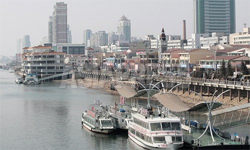 中国丹東市の河岸。高層ビルと遊覧船、そしてたくさんの観覧客がひしめきあう観光地の姿だ。