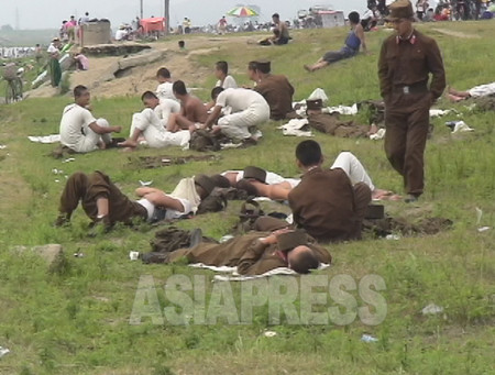 公園で休日を楽しむ住民の横で休憩中の兵士たち。「庶民より兵士の方が元気がない。腹を減らしているから」と撮影者のリ・ジュンは言う。（撮影：2006年8月清津市）