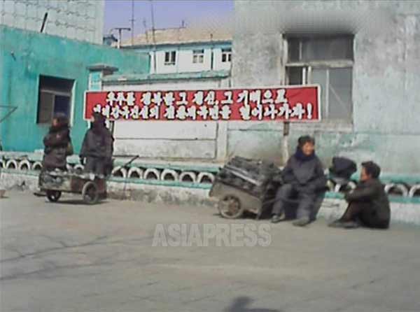 道端で練炭を売る女性たち。北朝鮮では「クモンタン（穴の空いた炭）」と呼ばれる。背後には「宇宙を征服したその精神・気迫で強盛国家建設の転換的局面を切り開いていこう！」というスローガンが見える。平城（ピョンソン）市、撮影 アジアプレス