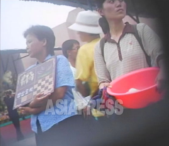 「食卓塗色」と書かれた紙を持って市場に立つ女性（左）。技術労働の求職だ。（2007年8月平壌（ピョンヤン）市楽浪（ランラン）市場　リ・ジュン撮影）