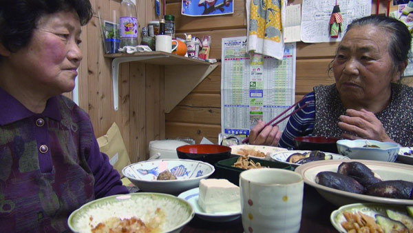 福島県伊達市の仮設住宅で。食卓には、避難先で借りた畑でつくった料理が並ぶ。とれたての新鮮な野菜のおいしさは忘れられないと古居監督は話す。（映画「飯舘村の母ちゃんたち　土とともに」より）