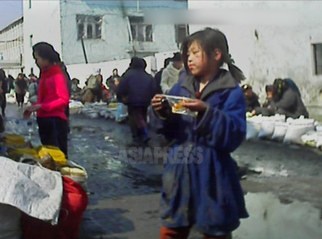 腹を空かせているのだろうか。少女のコチェビが、露天市場で食べ物をじっと見つめている。手には食べ物が少し入ったビニール袋が握られている。2013年3月平安南道平城(ピョンソン)市にて撮影アジアプレス。