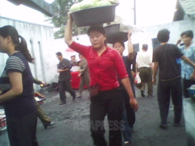 白菜かキャベツだろうか、売り物を入れた大たらいを運んでいる。頭に載せることを「イダ」といい、南北問わず朝鮮民族の習慣。男性はやらない。2010年6月平安南道にて撮影キム・ドンチョル（アジアプレス）