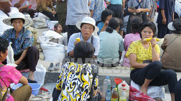 北朝鮮のごくありふれた市場の様子。ゆでトウモロコシを頬張りながら米や餅、パンなどを売っている。2008年8月平壌市郊外にて撮影チャン・ジョンギル（アジアプレス）