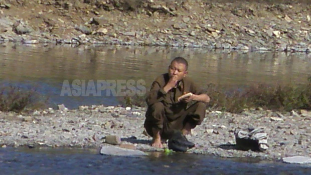 川辺で靴を脱いでトウモロコシをついばむ下級兵士。2008年9月平壌市郊外にて撮影チャン・ジョンギル（アジアプレス）