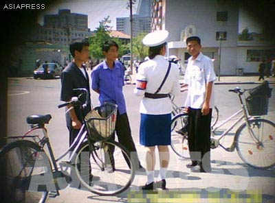 These three men were stopped at an intersection and reprimanded for riding their bicycles in an illicit manner. Usually, when detaining a citizen, the officer starts by confiscating his or her identification card. This move prevents resistance and ensures obedience most of the time. Citizens typically resort to the fastest and easiest way of resolving the problem: bribery. The "price" was around 500 won (12 U.S. cents) at the time this photo was taken. [Gu Gwang-ho, 2011]