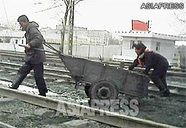 A man and woman pull a large handcart across railway tracks. The man has lashed his body to the cart with a rope so as to pull the cart more effectively. (South Pyongan Province. March, 2013)ASIAPRESS