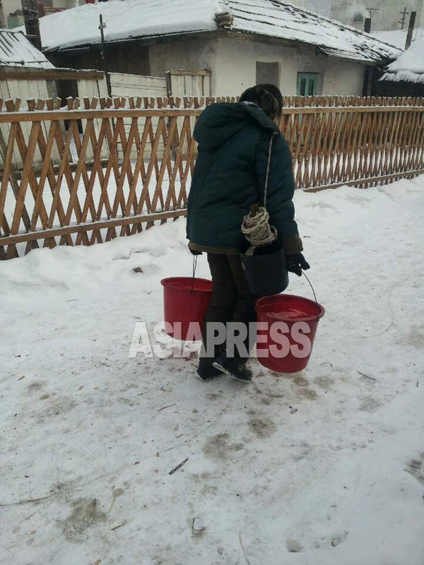 A woman is heading for her home with buckets of water from the public well. The biggest challenge is to secure water for living during winter in North Korea.  The river is frozen and there is no running water as the motor pump does not work because of insufficient power supply.  Residents collect money together to dig a well or break ice on river to draw water. (Taken by Mindulle/January 2015/ ASIAPRESS)